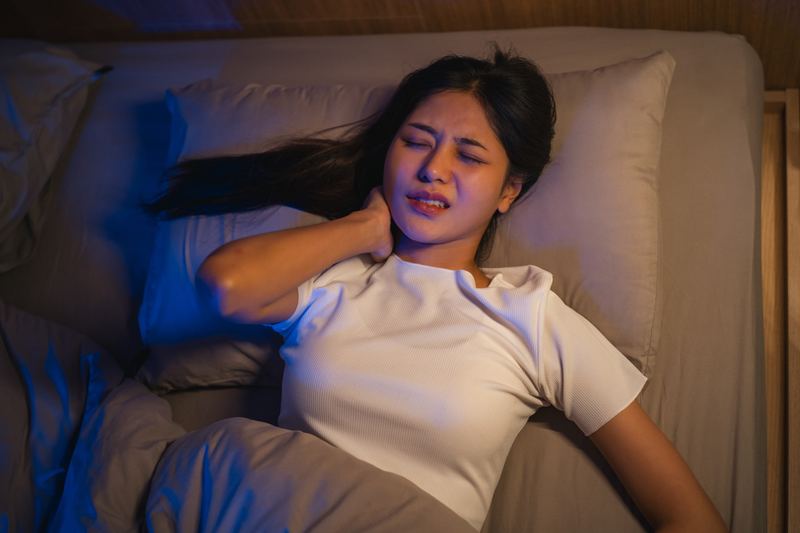 夜、ベッドで首肩の痛みに顔をしかめる女性